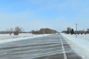 Snow blowing across the tree lined country highway on a bright winter day