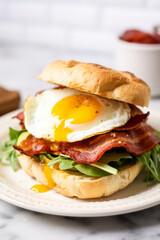 Bacon and egg breakfast sandwich in a white kitchen on the table with a runny yolk close up - 735398635