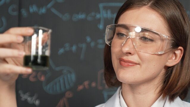 Closeup of young beautiful teacher focus on looking chemical liquid at laboratory. Skilled scientist doing an experiment by inspect colored solution while standing in front of blackboard. Erudition.