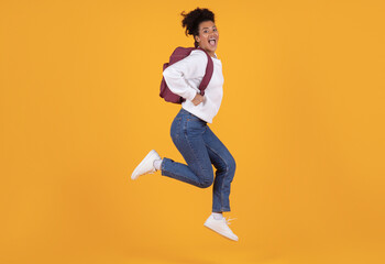 Fototapeta na wymiar Joyful young black woman wearing backpack jumping in mid-air against yellow background