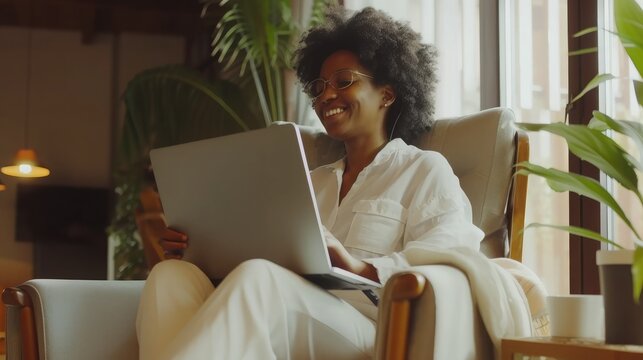 Image of cheerful african american woman using laptop while sitting on chair in living room