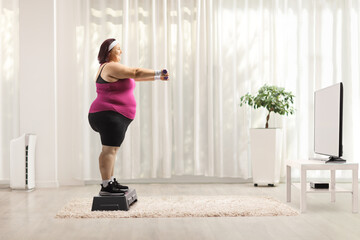 Fototapeta na wymiar Plus size woman exercising with dumbbels in front of tv