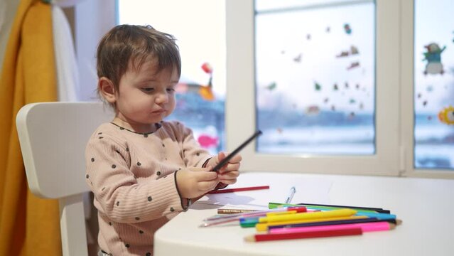 baby girl draws while sitting at a table by the window at home. happy family kid concept. baby daughter learns to draw with pencils on a sheet of paper indoors. development of fine dream motor skills