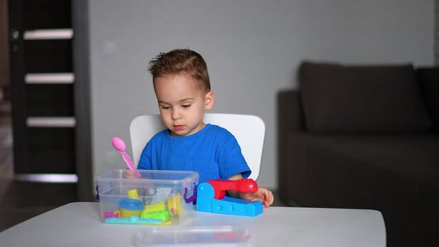 Beautiful dark-haired baby boy sitting at white desk. Cute kid pulls the toy from the plastic box.