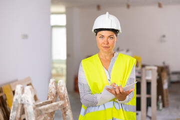 Woman foreman in a protective helmet and yellow vest checks the completed construction work on the drawing