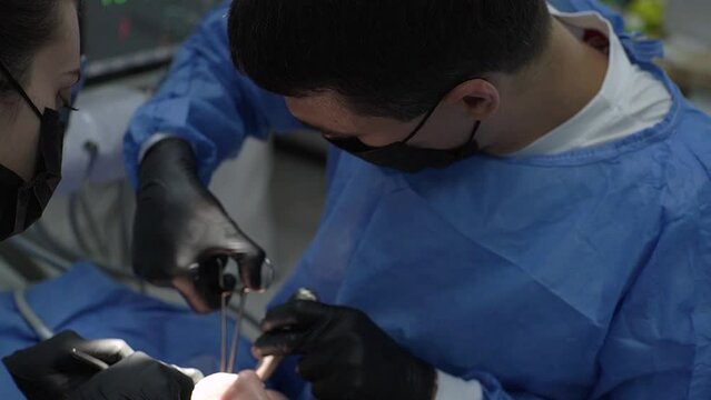 Close-up. surgeon's hands, black gloves, perform operation remove impacted wisdom tooth together assistant, nurse. Saliva ejector blood. Surgical suit, uniform. wound mouth sutured clamp. Sleep