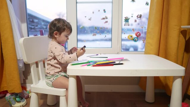 baby girl draws while sitting at a table by the window at home. happy family kid concept. baby daughter learns to draw with dream pencils on a sheet of paper indoors. development of fine motor skills