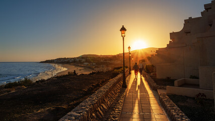 A couple of tourists walking along a walkway illuminated by vibrant sunlight at sunset on Costa Calma beach in the island of Lanzarote in Canaries