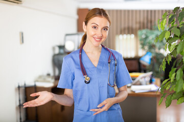 Polite smiling young woman therapist in blue uniform with stethoscope on her neck standing in medical clinic, inviting for consultation