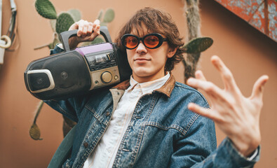 Young stylish male hipster wear in retro denim jacket 90s old fashioned style hold audio tape cassette player and showing gesture by fingers like rap singer.