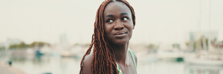 Woman with African braids wearing top looks at the yachts and ships standing on the pier in the...