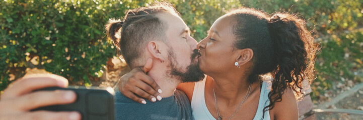 Happy smiling interracial couple kissing while sitting on bench and take selfi, Panorama