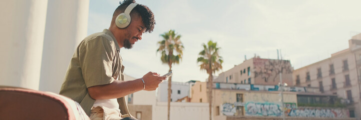 Young male student dressed in an olive color shirt sits outside next to the university, listens to music on headphones, selects tracks on his phone, Panorama