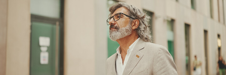 Panorama of mature businessman with beard in eyeglasses wearing gray jacket walks down the street past modern buildings