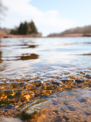 A very small wave in the shallow area of a coastal river. The wave created ripples in the water's surface. There are many small pebbles of multi colors just below the water. There is blue sky and sun.