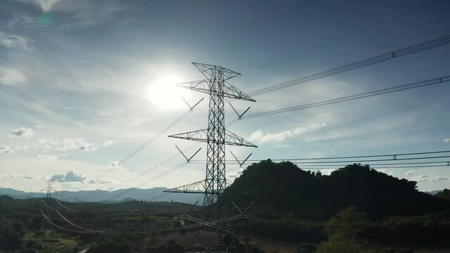 Backlit aerial view of a steel lattice transmission tower and high-voltage power lines over wooded hills in Pha Sing Subdistrict, Mueang Nan District, Nan Province, Thailand, electric utility grid 4K.