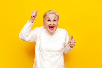 cheerful old grandmother in a white sweater wins and rejoices at success on a yellow isolated...