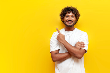 young indian man in white t-shirt pointing at copy space and smiling on yellow isolated background, curly guy showing and advertising empty space - obrazy, fototapety, plakaty