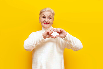 cheerful old grandmother in a white sweater shows a heart with her hands and smiles on a yellow...