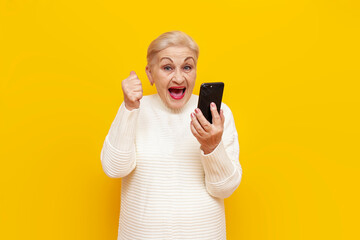 cheerful old grandmother in a white sweater using a smartphone wins and rejoices at success on a...