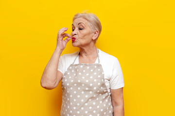 cheerful old grandmother housekeeper in an apron shows a bellissimo gesture on a yellow isolated background, an elderly woman housewife shows a sign of good taste - obrazy, fototapety, plakaty