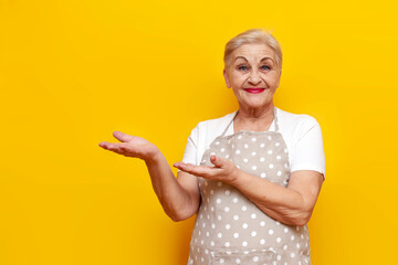 cheerful old grandmother housekeeper in apron points with hands at copy space on yellow isolated...