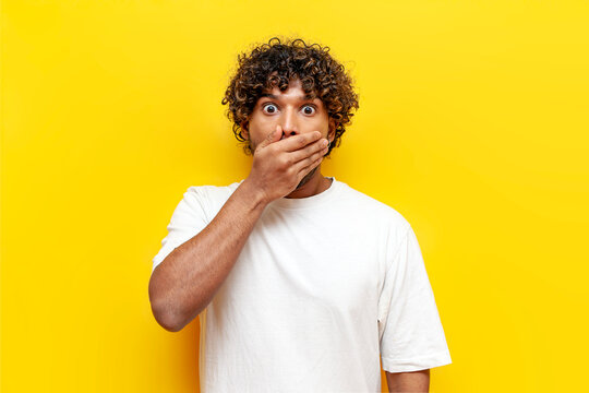 young shocked indian man in white t-shirt covering mouth with hand on yellow isolated background, curly surprised guy looking at camera with big eyes in amazement and fear