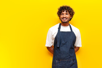 cheerful indian male waiter in an apron stands and smiles on a yellow isolated background, indian...