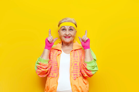 crazy funny old granny in sports colorful clothes points up on a yellow isolated background, elderly woman in youth hipster clothes shows and advertises copy space from above
