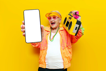 crazy cheerful grandmother with a tape recorder shows a blank screen of a smartphone on a yellow...