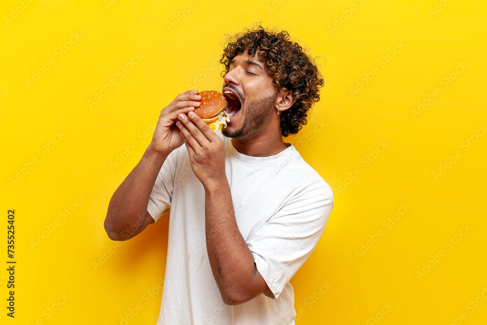 Wall mural young indian man eating a big tasty burger with his mouth wide open on a yellow isolated background, curly guy student eating and advertising fast food