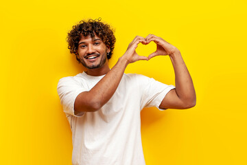 curly indian man in white t-shirt showing heart with hands on yellow isolated background, young...
