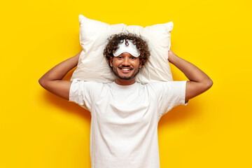 young cheerful indian man in pajamas and sleep mask lies on a soft pillow resting and smiling on a yellow isolated background, curly guy getting ready for bed and hugging a pillow - obrazy, fototapety, plakaty