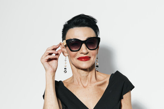 Elegant woman with red lipstick and black dress holding sunglasses to face, fashionable and chic look