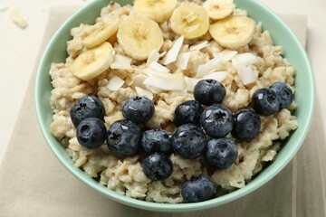 Tasty oatmeal with banana, blueberries, coconut flakes and honey served in bowl on beige table, closeup