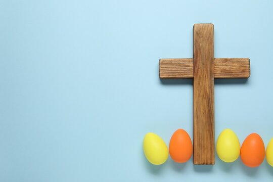 Wooden cross and painted Easter eggs on light blue background, flat lay. Space for text