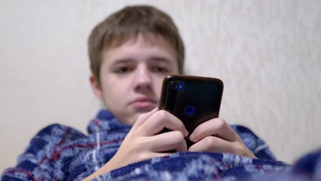 Teenager Browsing Social Networks on a Smartphone While Lying on a Bed in a Room. Close up. Boy in pajamas reads messages, chats, and scrolls through a video in a mobile app before going to bed. Blur.