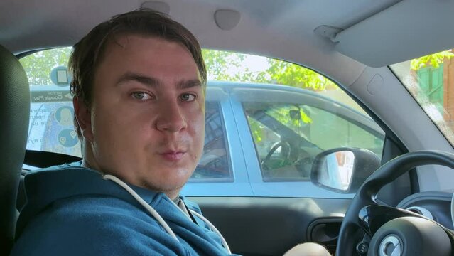 plump, tired man in a hoodie sits behind the wheel of a parked car, talks to a passenger, shakes his head with displeasure, shares his problems and experiences Chatty taxi driver complains about life