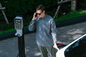 Young man travel with EV electric car charging in green sustainable city outdoor garden in summer...