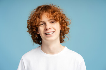 Portrait of smiling confident boy, curly red haired teenager with dental braces on teeth looking at camera isolated on blue background. Health care, treatment, orthodontic concept