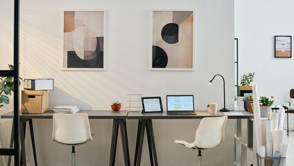 No people shot of part of modern office interior with laptop, digital tablet, pile of documents on desks and paintings on walls, copy space