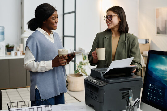 Young Caucasian secretary holding cup of hot drink printing document and chatting with her African American colleague