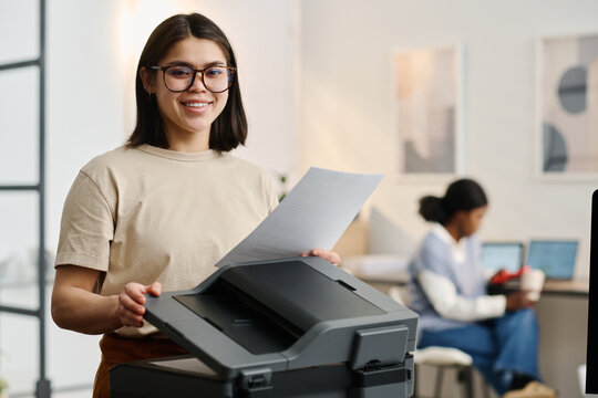Selective focus medium portrait of cheerful gen Z female employee wearing eyeglasses smiling at camera while scanning documents in modern office