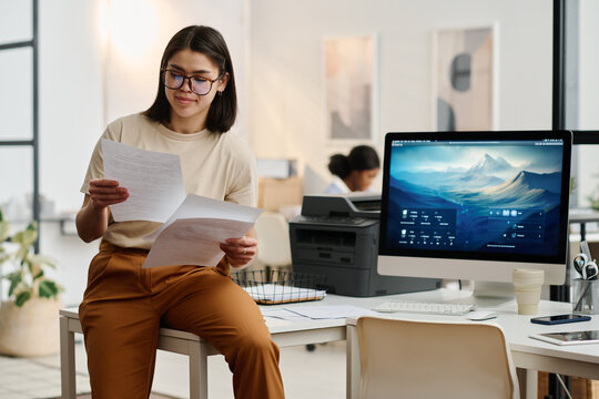 Medium long shot of young gen Z female CEO sitting on table in modern office looking through documents, copy space