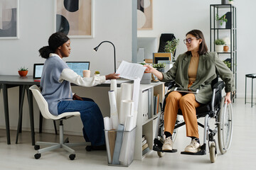 Long shot of young Caucasian woman in wheelchair and her African American colleague working in modern office
