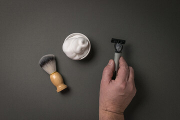 Shaving foam, a shaving brush and a man's hand with a razor on a gray background.