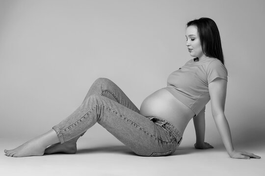 Black and white photo of young pretty pregnant woman in t-shirt and jeans sit on the floor on gray background. Female with belly exposed.