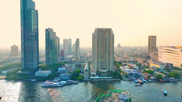 4K - Bangkok Thailand: Aerial view, Sunset paints the downtown with golden hues, as a drone navigates the labyrinth of towering buildings lining the peaceful river. City life concept.
