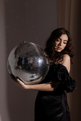 A young Caucasian woman in an elegant black off-shoulder dress holds a large, gleaming disco ball....