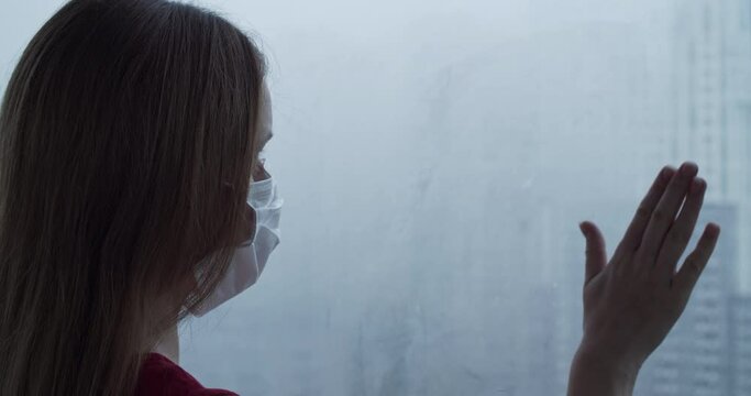 Woman with mask gazes out foggy window, contemplative mood amidst urban, reflecting resilience and solitude during challenging times. Evoking feelings of isolation yet hope during social distancing.